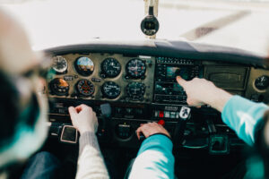 A pilot and a passenger sit in the cockpit of a small aircraft pointing at the tachometer systems