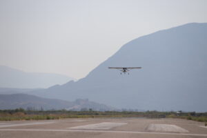 Small single-engine airplane taking off from a rural runway with mountains in the background on a hazy day.