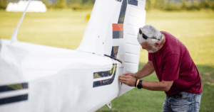 Man inspecting the tail section of a small white airplane on a grassy airfield.