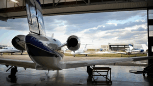 Private jet parked inside an aircraft hangar with other small planes visible outside under a cloudy sky.